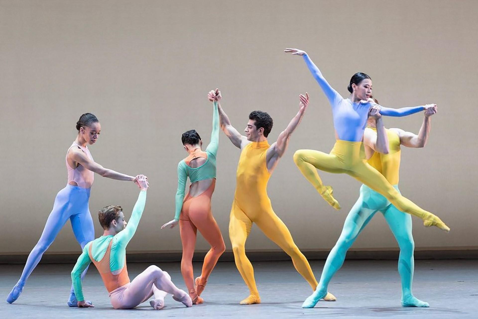 Six ballet dancers in pastel and bright colored costumes perform on stage; two are holding hands as one leaps, while others pose and interact gracefully under soft lighting.