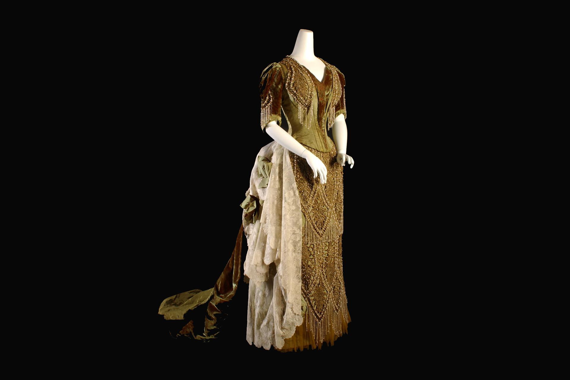An elegant vintage evening gown with short sleeves, beaded details, a fitted bodice, and a long flowing train adorned with lace and fabric draping, displayed on a mannequin against a black background.
