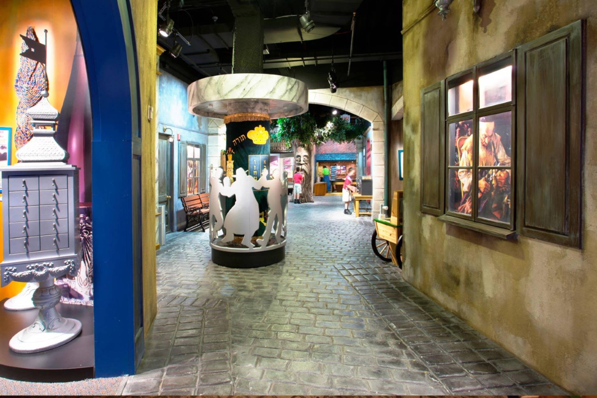 A colorful, museum-like exhibit with cobblestone floors, painted walls, artwork displays, and decorative elements, including a round structure with silhouettes of people dancing in the center.