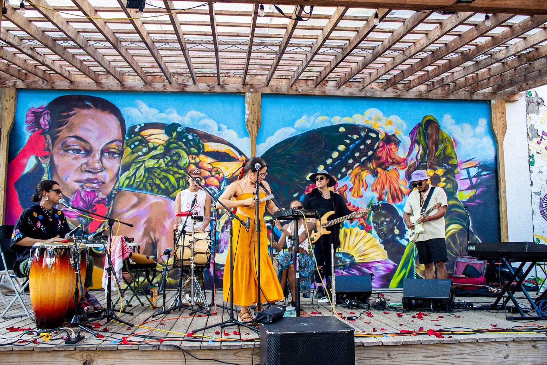 A band performs on an outdoor stage with colorful murals behind them. The musicians play various instruments, including guitar, keyboard, and drums. The stage is scattered with flower petals.