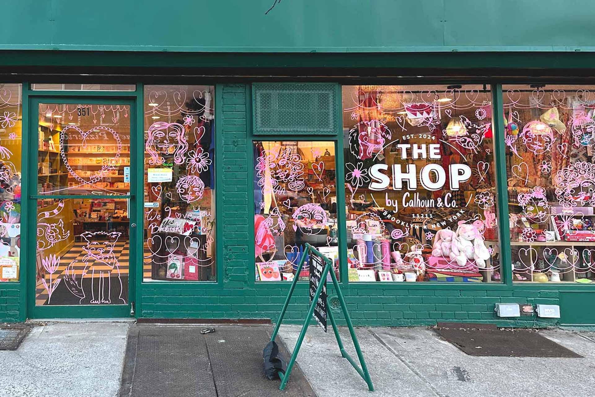 Storefront of "The Shop by Calhoun & Co." with green trim. Windows display pink and white whimsical illustrations and various colorful products like plush toys and home decor. A green sandwich board stands on the sidewalk.