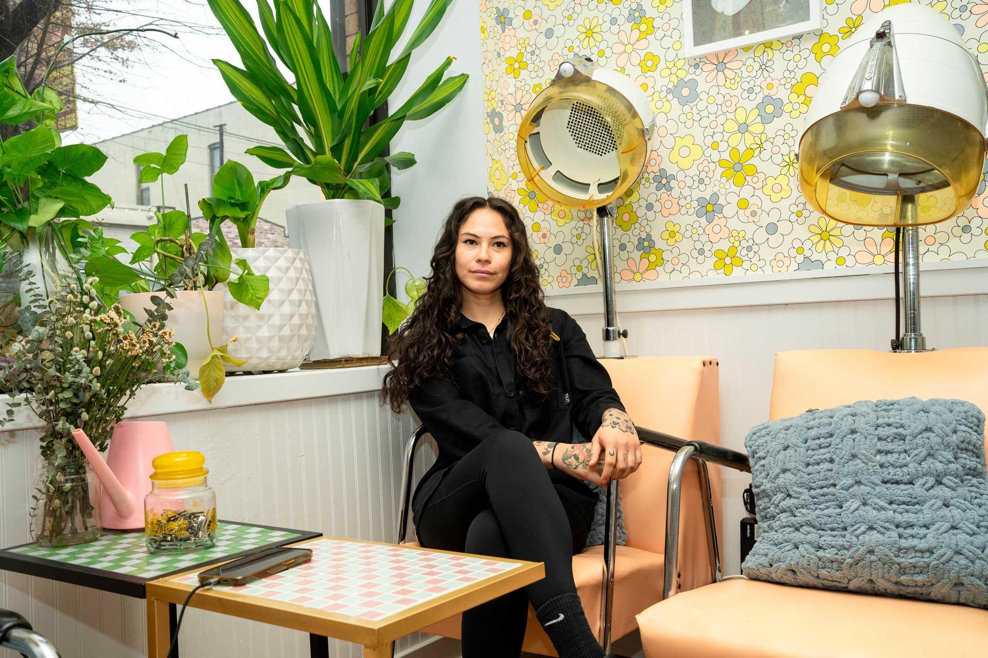 Yara Herrera, with long curly hair, sits on a peach-colored chair at Queen of Swords, a retro-style salon with vintage hair dryers, potted plants, and colorful floral wallpaper. She is wearing a black outfit and looking at the camera.