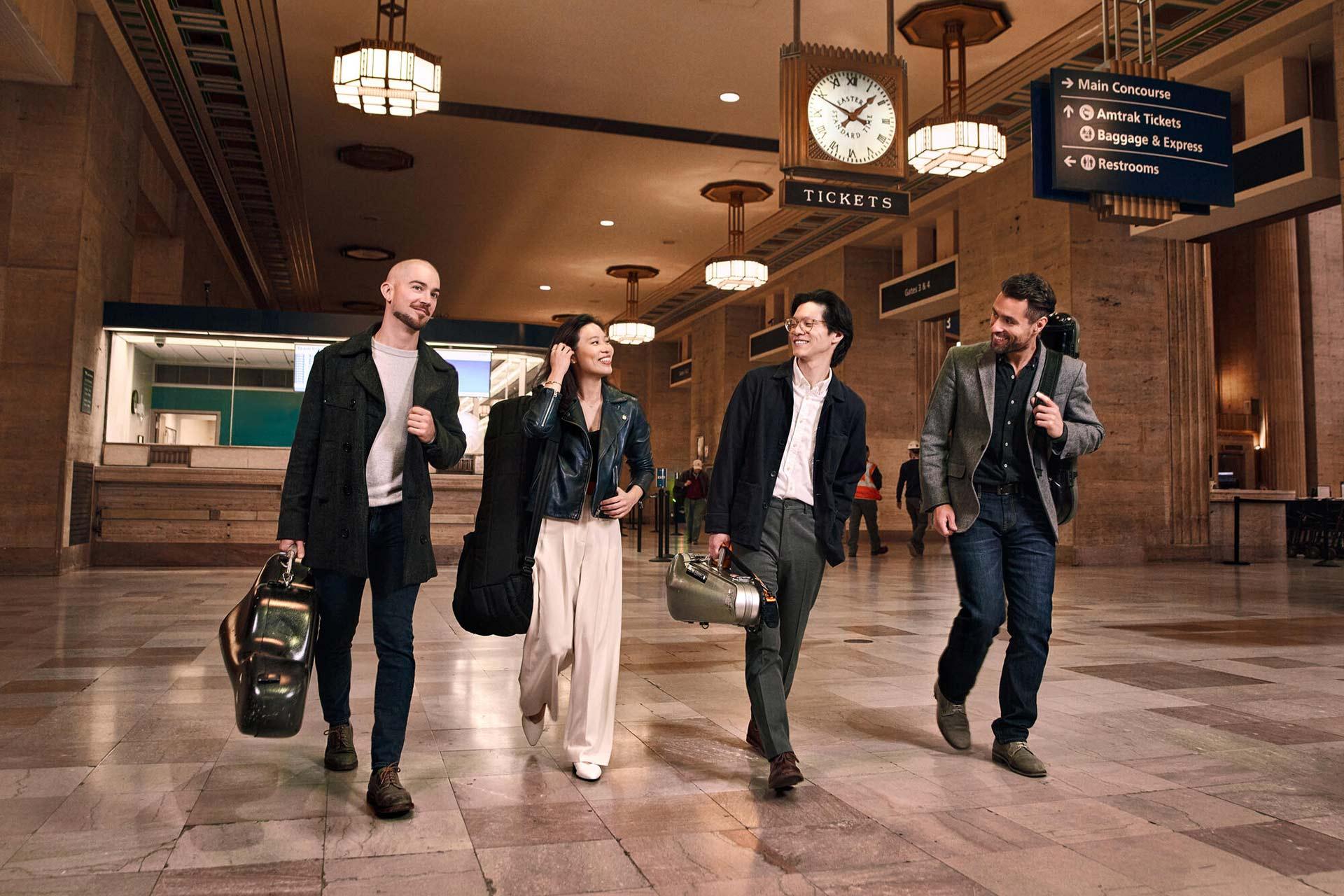 Four people, dressed in semi-formal attire and carrying instrument cases, walk together and smile inside a large, ornate train station with high ceilings and a prominent clock overhead.
