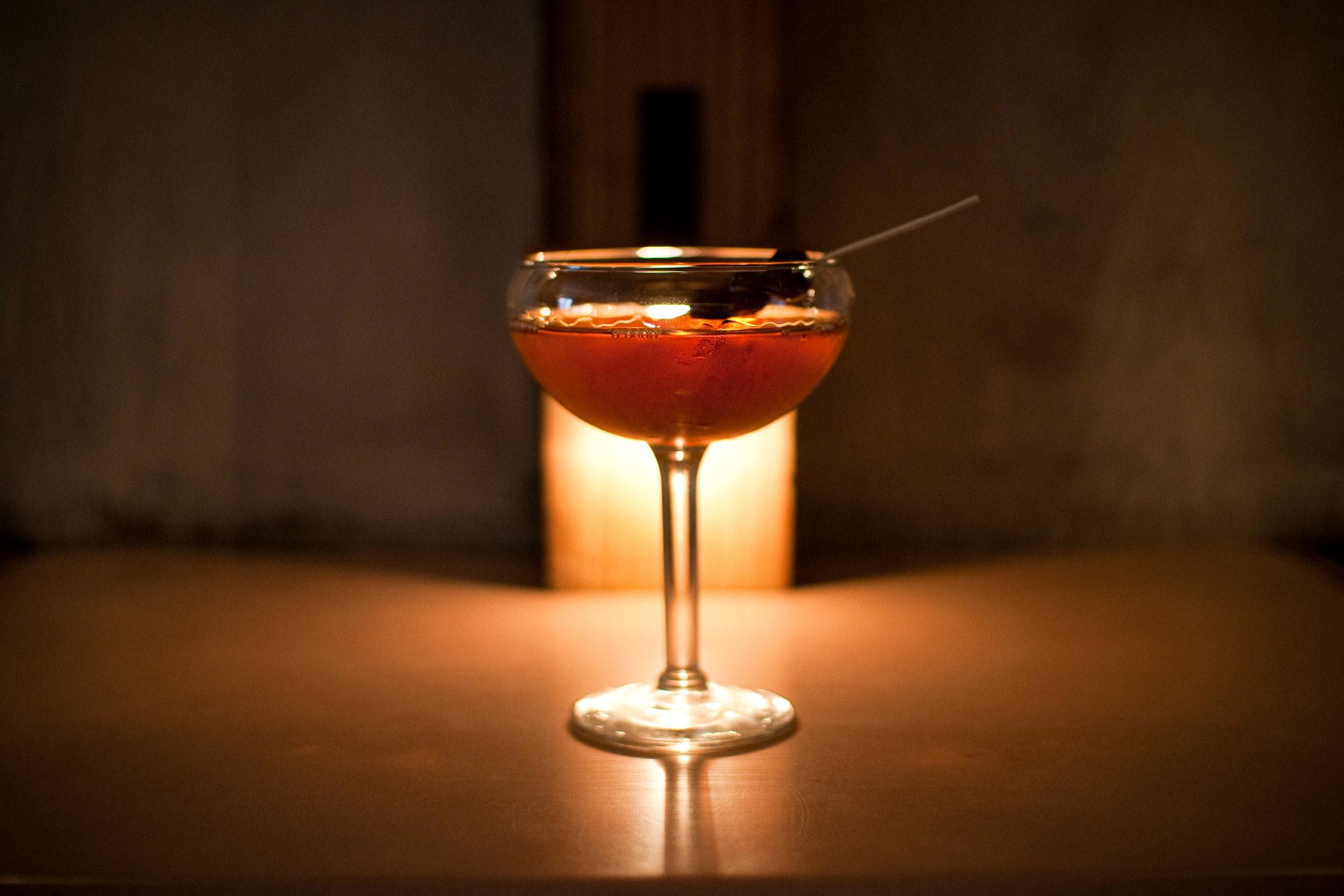 A cocktail in a coup glass sits on a table with a dim, warm light in the background. The drink is amber-colored and garnished with a dark cherry on a skewer. The setting appears cozy and intimate.