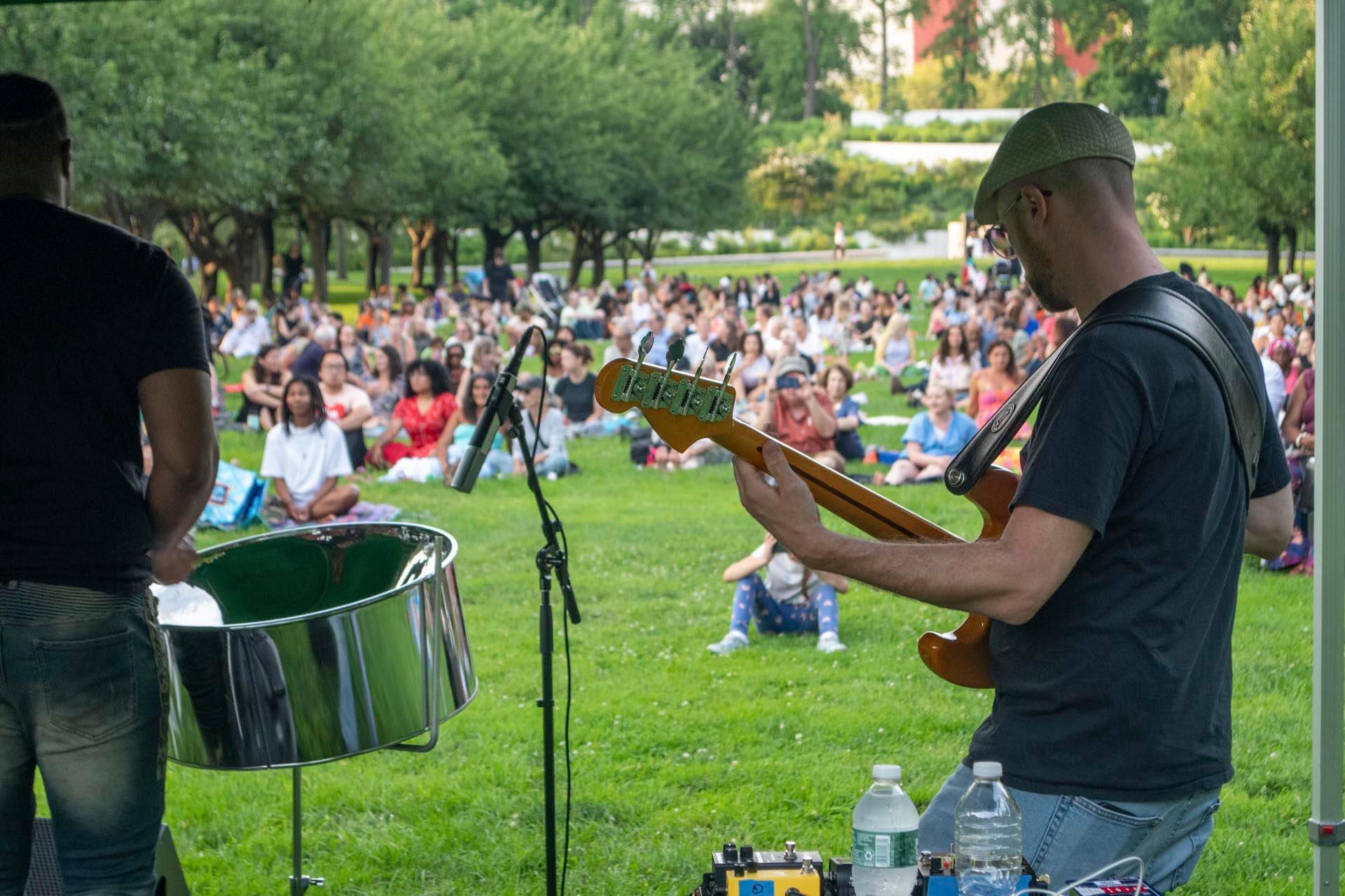 A musician plays electric bass onstage at an outdoor concert in a park, while a large seated audience watches on the grass in the background. Trees and greenery surround the event.