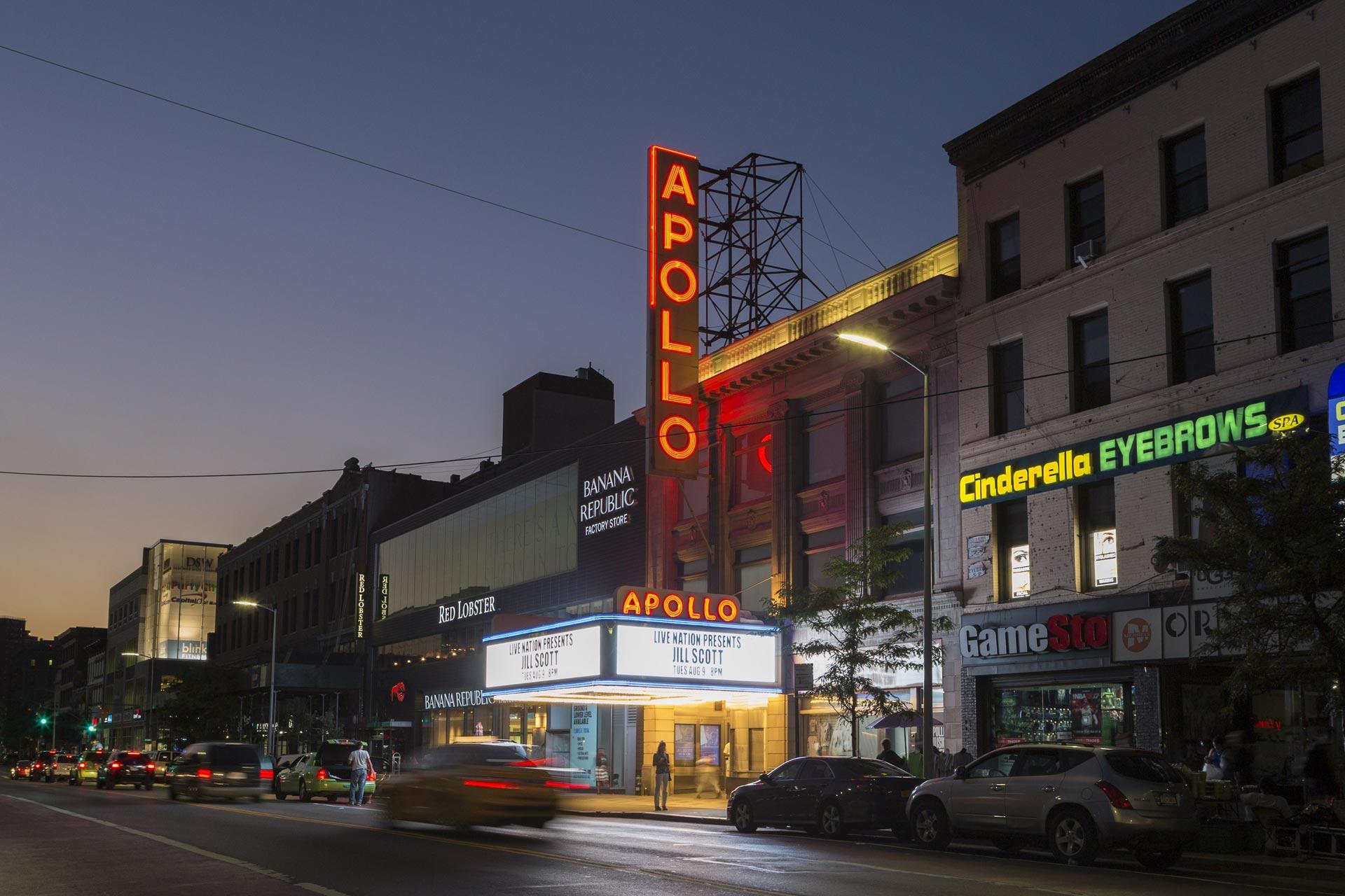 The Apollo Theater at dusk, with its bright vertical neon sign and marquee lit up. Nearby, there are storefronts for Banana Republic, GameStop, and Cinderella Eyebrows, with cars parked along the street.