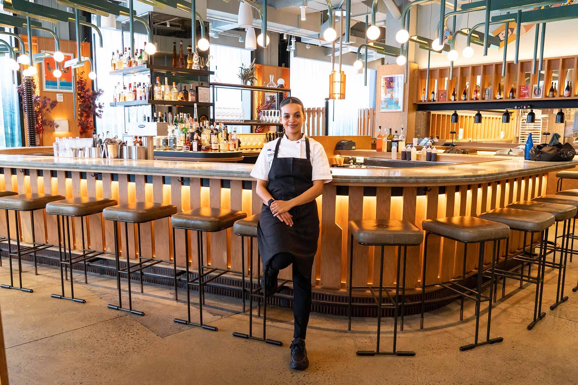 A chef in a black apron stands confidently in front of a modern, well-lit bar with wooden accents, high stools, and shelves stocked with bottles, giving the restaurant a welcoming atmosphere.