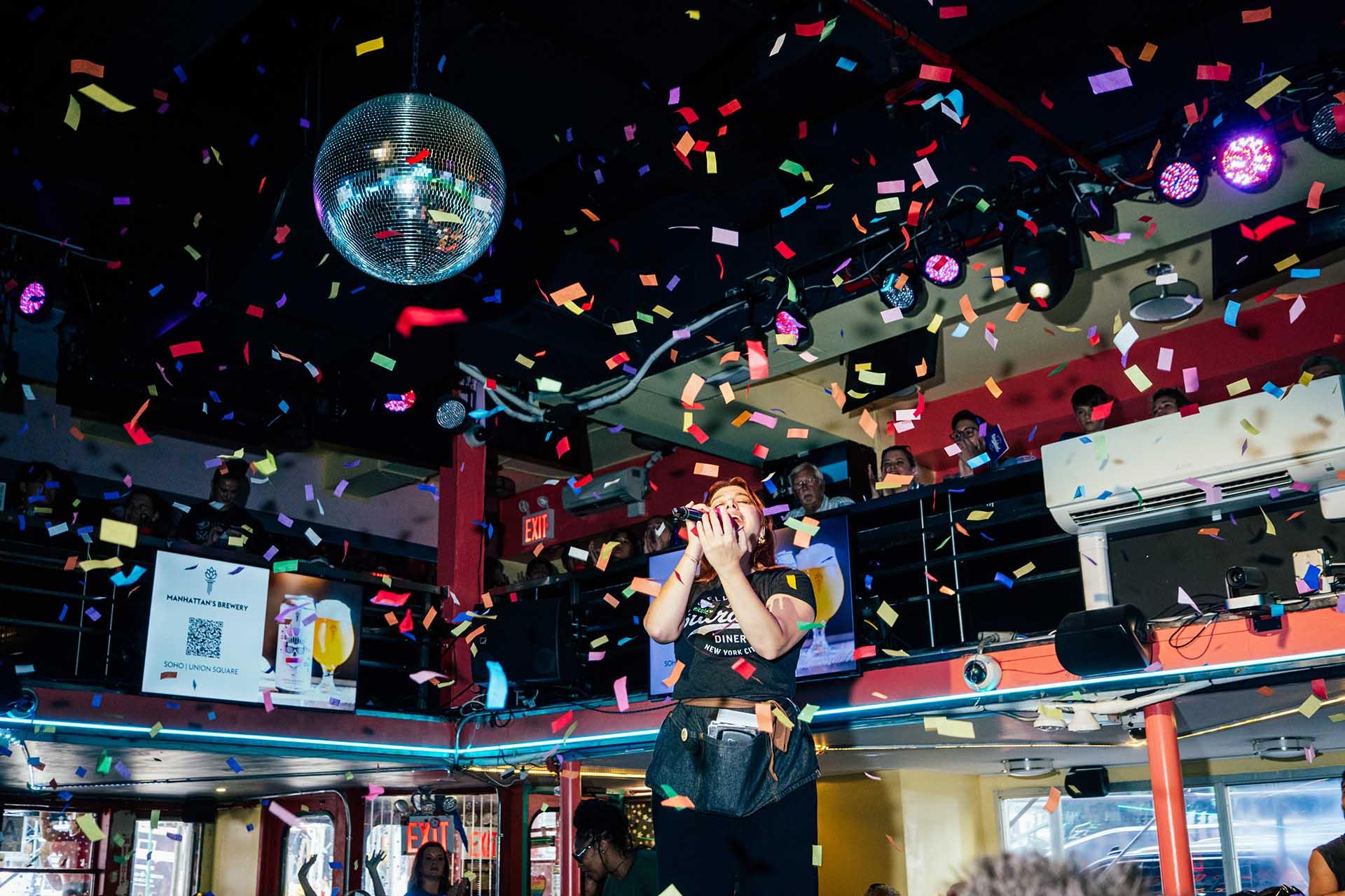A person stands on a stage in a lively bar, surrounded by colorful confetti falling from above, with a disco ball and audience watching from a balcony in the background.