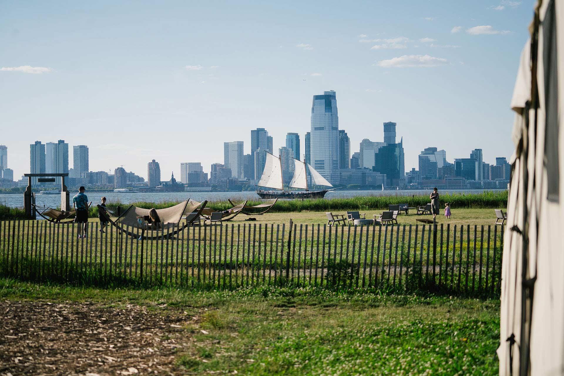 A grassy park with hammocks and people relaxing, bordered by a wooden fence. A sailboat is on a nearby body of water, with a city skyline featuring tall buildings in the background under a clear sky.