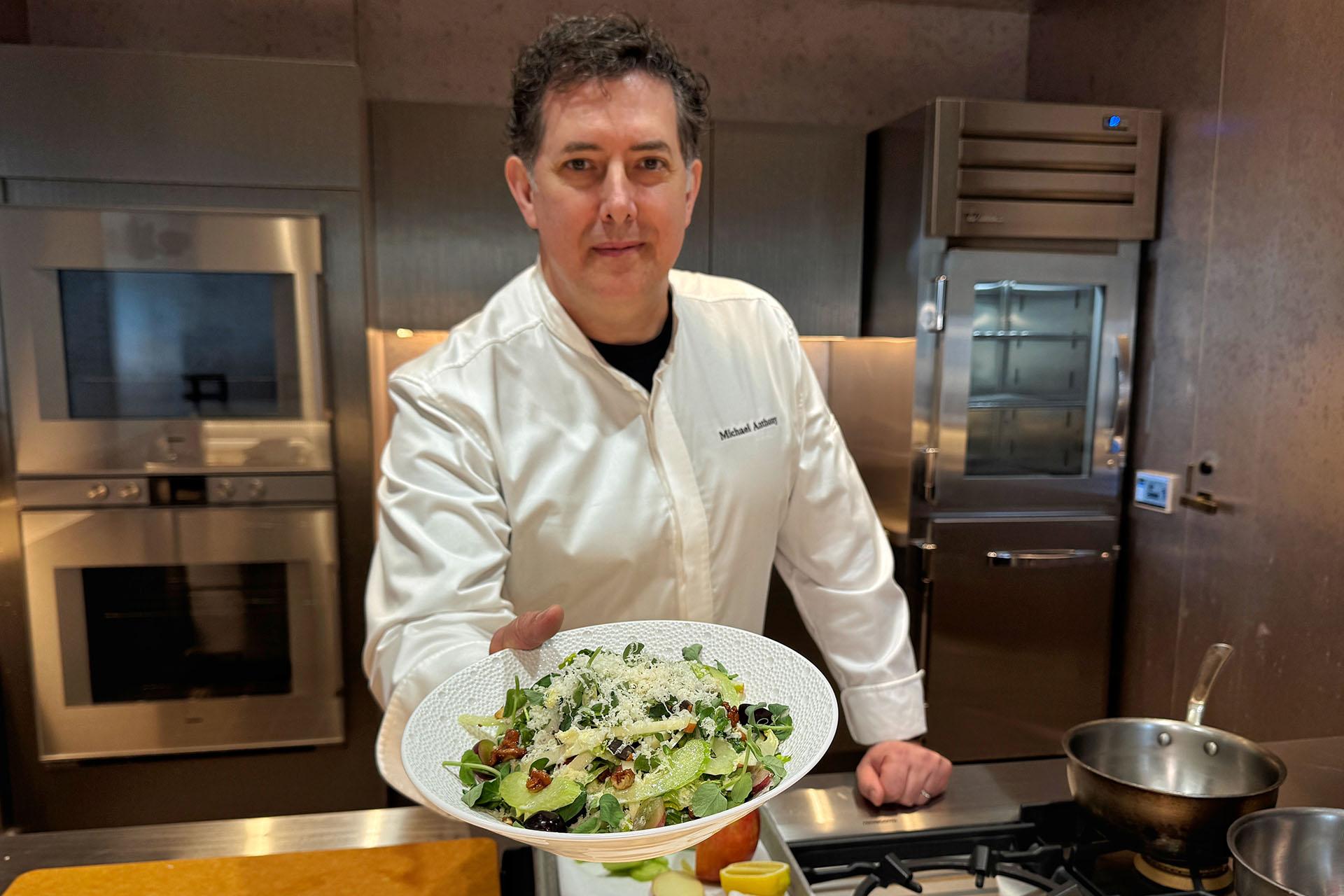 A chef in a white coat presents a fresh salad topped with grated cheese, greens, and olives in a white bowl over a kitchen counter with various ingredients.