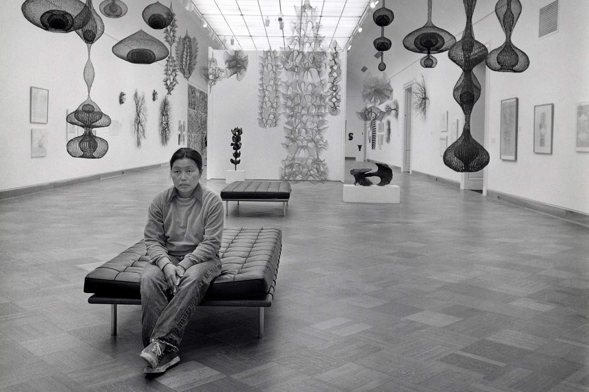 A person sits alone on a bench in a modern art gallery with intricate hanging sculptures and artwork displayed on white walls. The space is bright with a patterned wooden floor and a high ceiling.