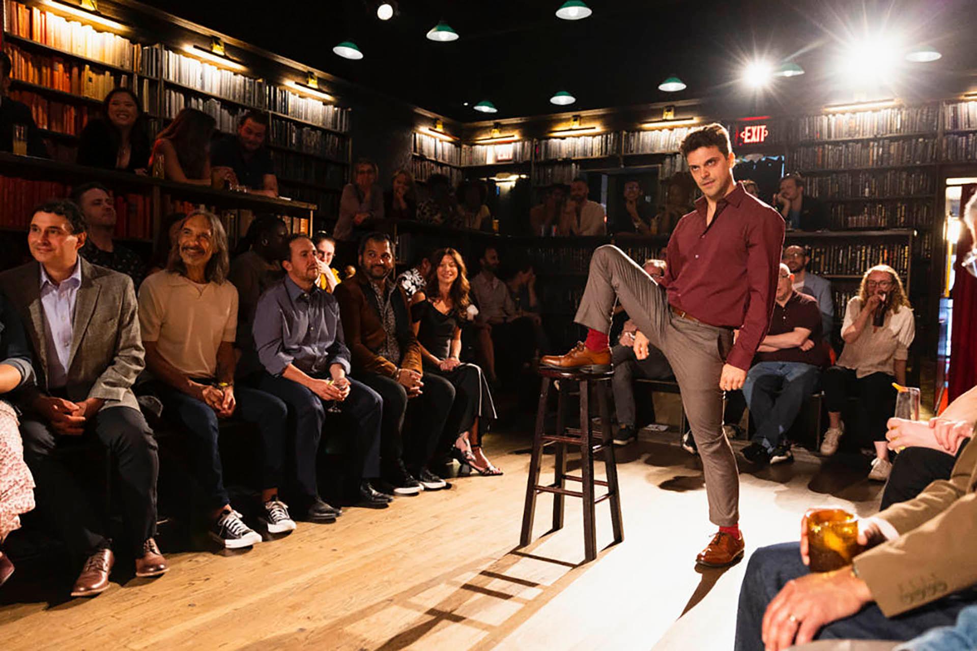 A man stands confidently with one foot on a stool in the center of a room, surrounded by an audience seated along the walls. The room has a warm ambiance, with bookshelves lining the walls and soft overhead lighting.
