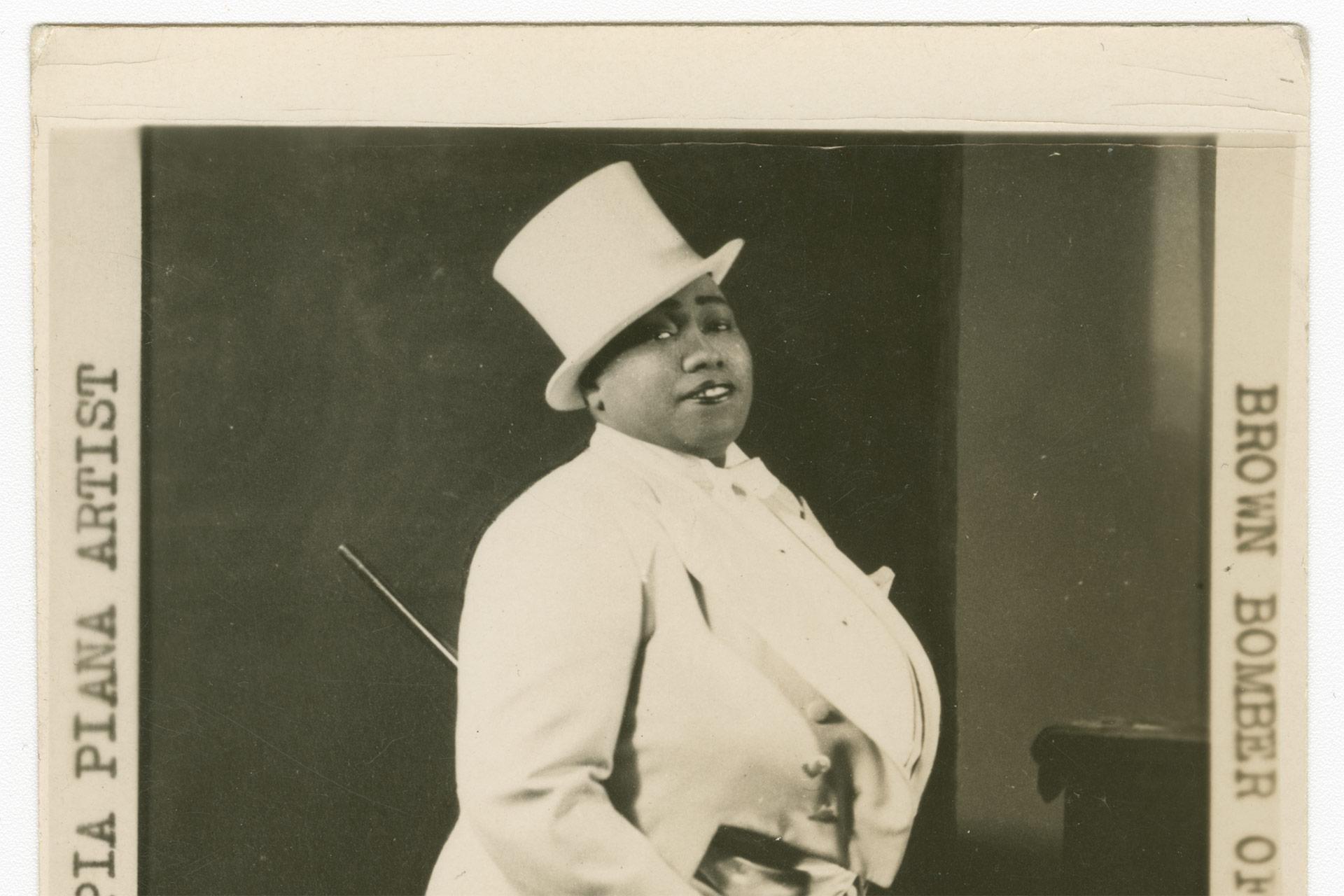 A person wearing a white tuxedo and top hat poses confidently with a cane, standing against a plain background in a vintage black-and-white photograph. Partial typewritten text is visible along the edges.