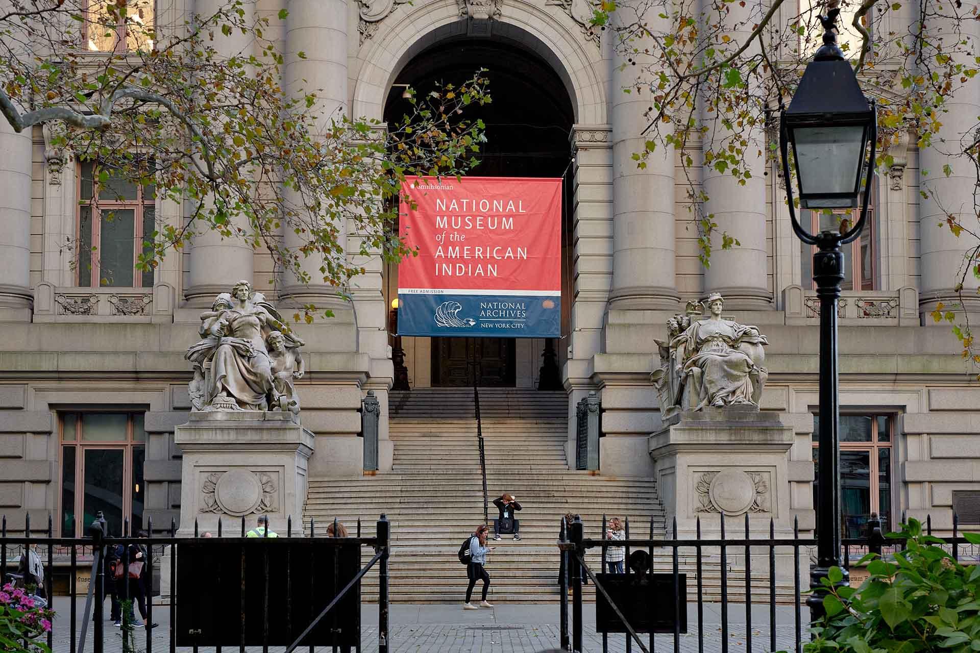 The image shows the entrance of the National Museum of the American Indian. It features grand steps, ornate statues on either side, and a large red banner above the entrance. Leafy trees and a classic lamppost frame the scene.
