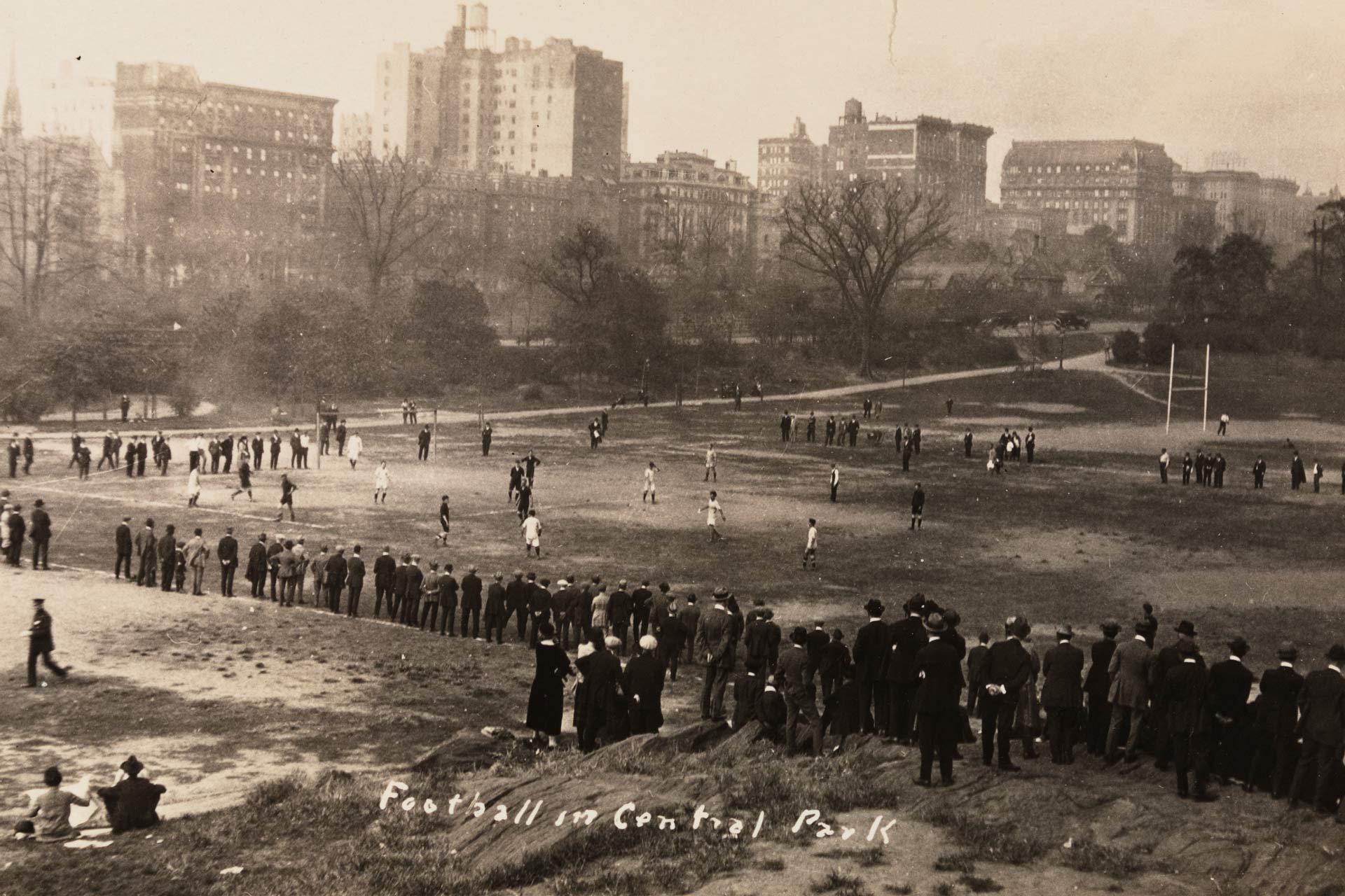 Black-and-white photo of a large group of people watching a soccer game in Central Park, New York City, with tall buildings and bare trees in the background. Spectators are gathered around the field.