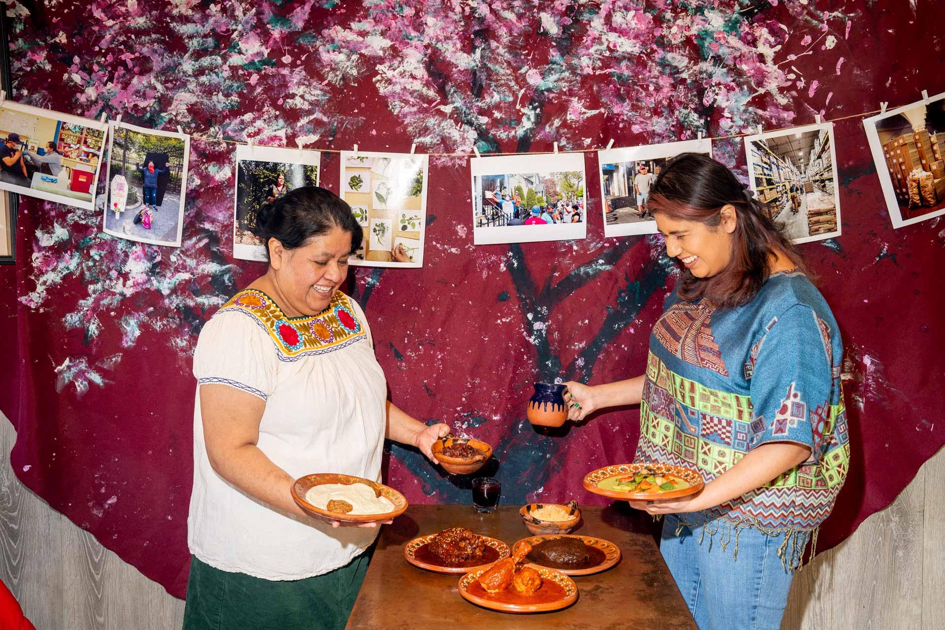 Natalia Mendez and her daughater Carolina Saavedra in colorful traditional clothing smile and serve food from a table covered with various Mexican mole dishes.