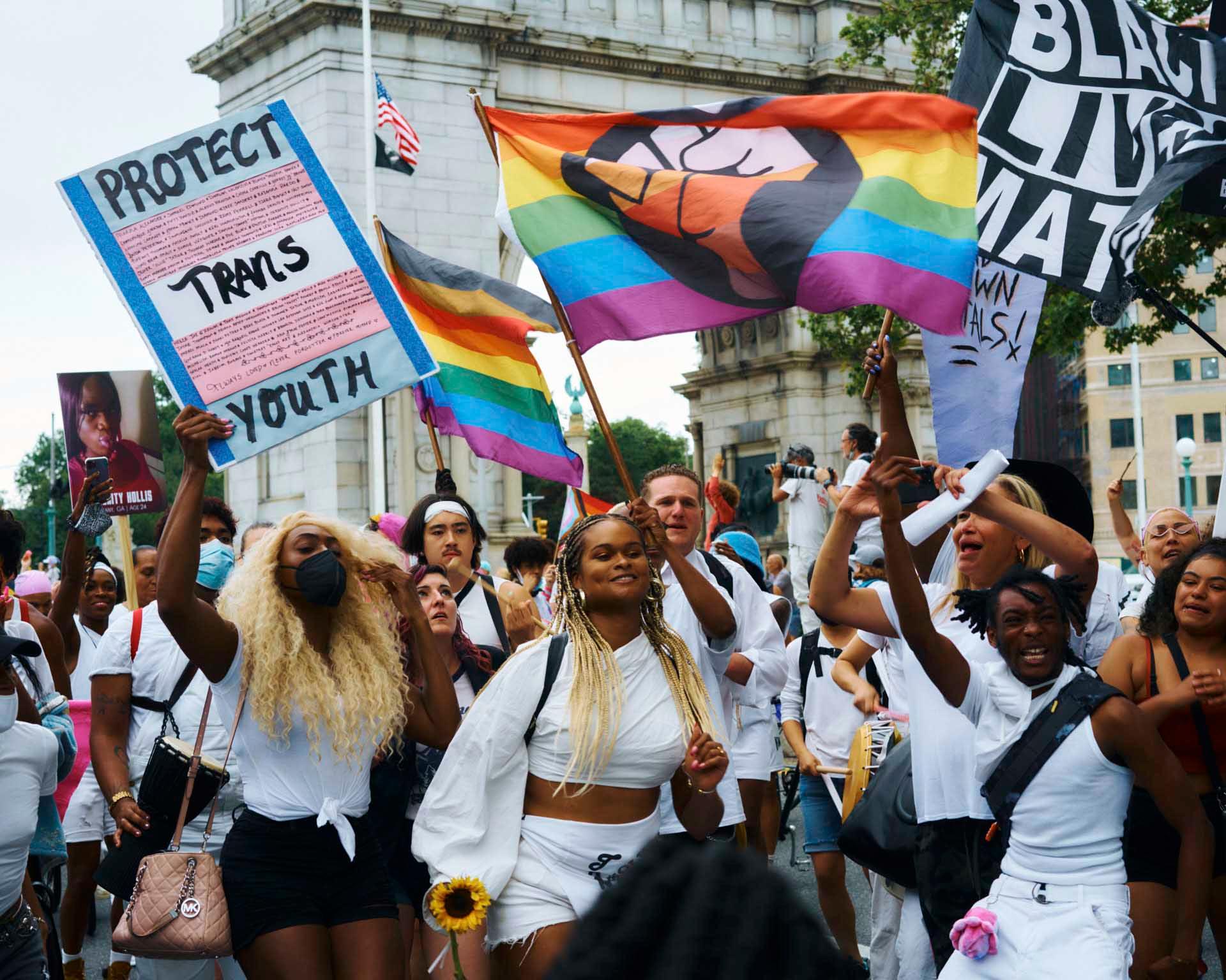 A large crowd of people, many dressed in white, gather on a city street in front of Barclays Center. A transgender pride flag is visible amid the crowd. Some individuals hold cameras, capturing the event.